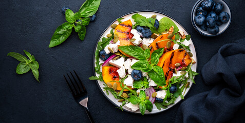 Gourmet salad with grilled peaches, blueberries, red onion, feta cheese, basil and mixed herbs, black table background, top view
