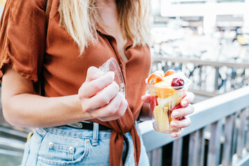 unrecognizable caucasian woman opening a cup of sliced fresh fruits. She is in the street