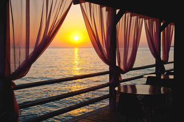 Obraz premium A beautiful image of a sunset over a calm sea from the curtains of a restaurant on the beach.