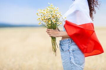 Close up of woman covered with  flag of Poland holding bouquet of daisies in wheat field. Polish...