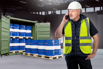 Man is logistician in hangar. Sea container with barrels. Logistician chemical enterprise. Pallets with chemical factory products. Man logistician speaks on phone. Warehouse contractor in yellow vest