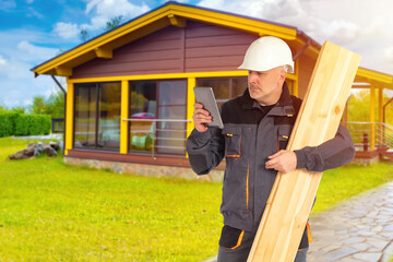 Man builds country house. Engineer is holding boards and tablet. Builder near wooden cottage. Man builder stands on lawn. Guy is building country house. Parquet board in hand of builder © Grispb