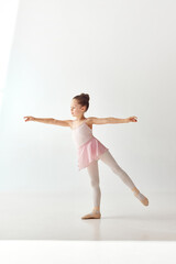 Portrait of small pretty school bun ballerina dancer girl in rose tutu ballet dress, white legging standing posing hold hands and leg up on white background. © Lustre Art Group 