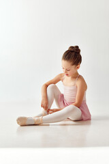 Portrait of small adorable preschool ballerina dancer girl in rose tutu ballet dress, white legging tying pointe against on white studio background.