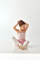 Portrait of small adorable ballerina dancer girl correct her hair bundle in rose tutu ballet dress, in pointe, white legging against on white studio background. © Lustre Art Group 
