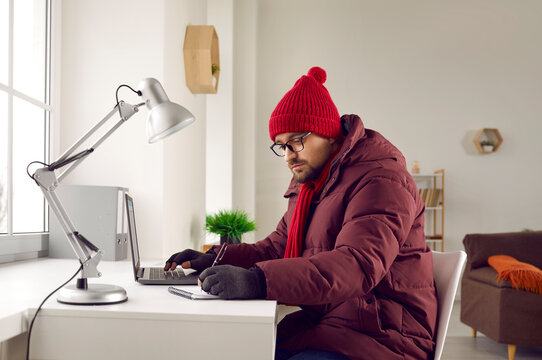 Freezing man in warm jacket and gloves working at computer in office. Unhappy warmly dressed businessman freezing in office since heating problems or power crisis at cold season