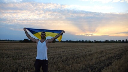 Young man walking with national blue-yellow banner on barley meadow at sunrise. Ukrainian guy going...