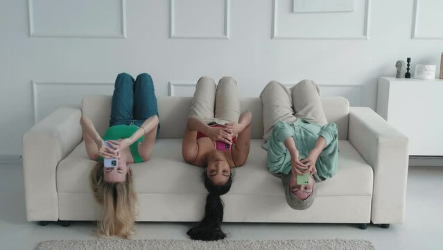 Multiracial girls friends lying on sofa with legs feet up, holding smartphone ignoring friends chatting online in messenger mobile phone addicted women, internet problem modern wireless tech usage