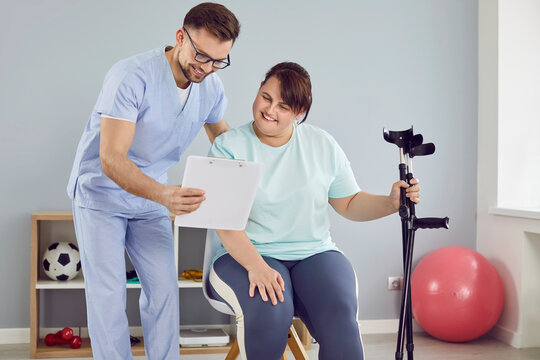 Cheerful friendly nurse showing fat young patient with crutches a report file with appointment in medical clinic. Physiotherapist or health care worker helping overweight woman in rehab and treatment