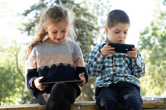 Children use gadgets while sitting in the park outdoors, games on smartphone and tablet