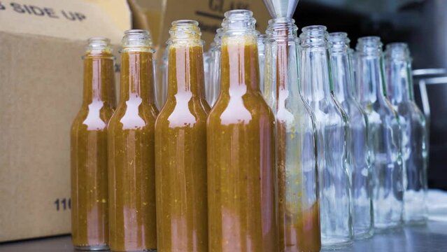 Filling up Cayenne chilli sauce into glass bottles with metal funnel and boxes background. Packaging production process in factory, USA.