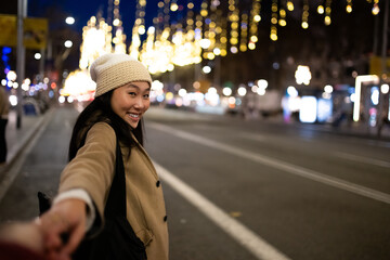 Beautiful woman holding hand in the street with christmas lights. Cheerful lady with warm hat looking at camera in winter. - obrazy, fototapety, plakaty