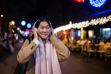 Young cheerful woman wearing headphones with christmas lights behind. Smiling girl listening to...