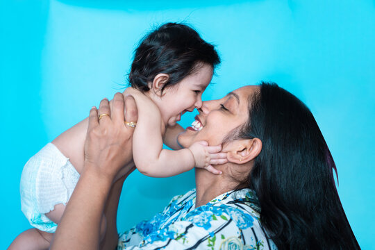 Smiling Indian Mother Playing With Her Six Months Cute Little Baby In Diaper Isolated Over Blue Background. Happy Family. Asian Mom With Infant Child Having Fun Lift In Air, Motherhood. Copy Space.