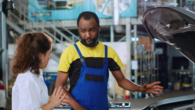 Certified worker in repair shop showing client damages found inside car after inspection. African american professional telling woman vehicle diagnostic after checking for defective parts