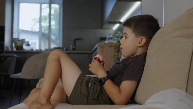 Side view of a caucasian boy wearing t-shirt eating red apple sitting on sofa at home, spending free spare time in living room. People lounge concept