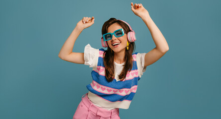 Joyful young woman in headphones and trendy glasses enjoying music while dancing on blue background