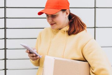 Young woman in red hat picks up parcel from automatic post office machine, Courier standing with phone and small box. Concept of fast delivery to automatic self lockers
