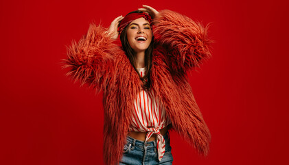 Attractive young hipster woman in fluffy coat looking at camera and smiling on red background