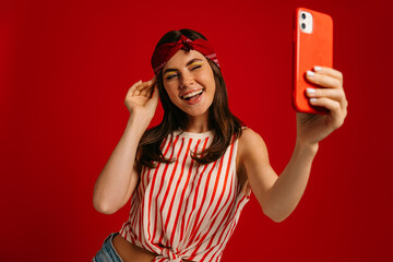Happy young hipster woman making selfie by mart phone and winking while standing on red background