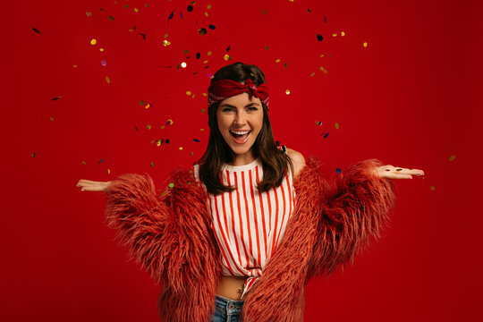 Joyful young hipster woman in fluffy coat throwing colorful confetti against red background