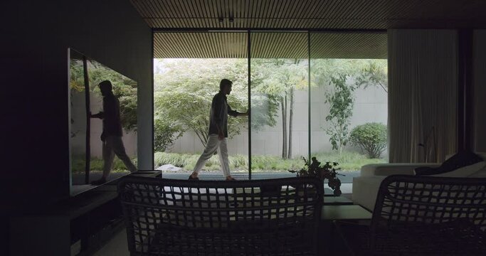 Young man open glass sliding door at modern cottage house. Modern vacation home with stylish furniture, contemporary interior and panoramic window with garden view.
