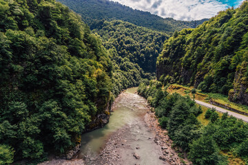 Beautiful landscape of mountains with green trees, roads along a mountain river on a sunny day with a cloudy sky.