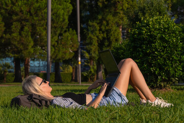 Smiling student uses a laptop lying on the grass. Young woman is lying with a laptop in a park on a sunny day.