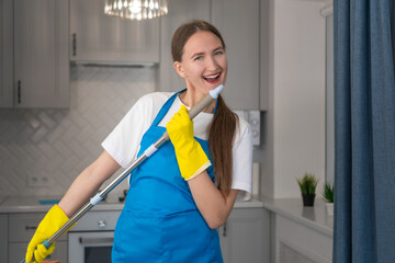 Happy young professional woman from a cleaning company cleans the house, sings into a mop as into a microphone, has fun.Housewife in an apron enjoys household chores, creatively engaged in cleaning - Powered by Adobe