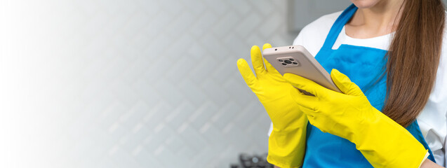 Close-up of a woman in yellow rubber gloves and a blue apron holding a smartphone and reading the news in the kitchen interior. Application and blog for social networks, house cleaning. Banner