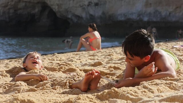 Child, tickling sibling on the beach on the feet with feather, kid cover in sand, smiling, laughing, enjoying some fun