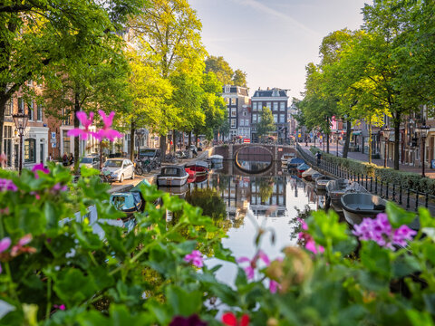 This photo showcases a peaceful Amsterdam canal scene. It includes an arched bridge, trees, boats docked in the canal and canal houses. Taken on a calm Sunday morning, the image reflects the canal's s