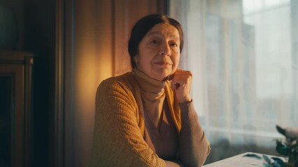 Pensive senior lady looking out window turning to camera with kind smile on face