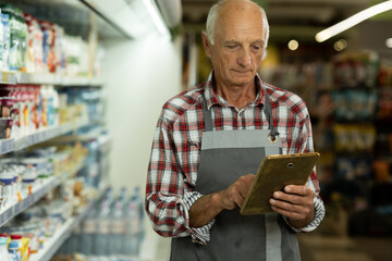 Elderly man using a digital tablet at supermarket, portrait of old male worker standing in supermarket and typing on tablet. Retail concept.