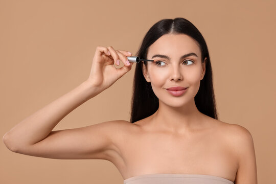Beautiful woman applying serum onto her eyelashes on beige background. Cosmetic product