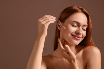 Fototapeta premium Beautiful young woman applying cosmetic serum onto her face on brown background, space for text