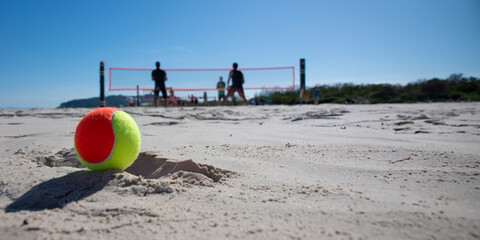 Doubles beach tennis game on the beach with blue sky and beach tennis ball in the foreground.