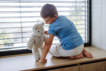 Diabetic boy with a continuous glucose monitor sitting by the window, showing his stuffed teddy...