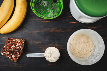 Shaker and scoop portion of whey, soy protein powder, protein chocolate bar and bananas on a dark wooden board, top view. bodybuilding food supplements, sports nutrition