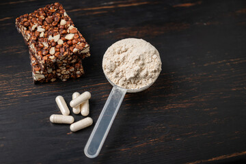 Scoop of whey or soy protein powder, white capsules of amino acids, vitamins, creatine, protein chocolate bar, bodybuilding food supplements, sports nutrition on a dark wooden board