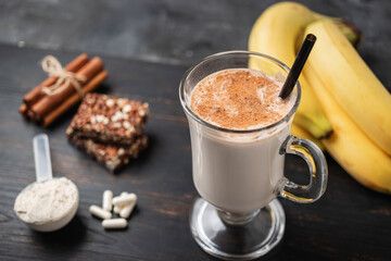 Glass of protein milkshake drink with straw and scoop of whey protein powder, white capsules of amino acids, vitamins, protein bar and bananas on a dark wooden board, bodybuilding food supplements