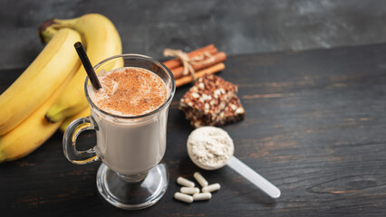 Glass of protein milkshake drink with straw and scoop of whey protein powder, white capsules of amino acids, vitamins, protein bar and bananas on a dark wooden board, bodybuilding food supplements