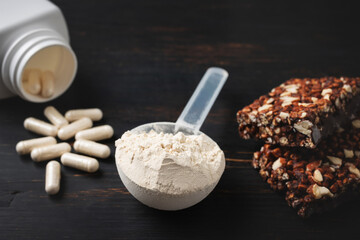 Scoop of whey or soy protein powder, white capsules of amino acids, vitamins, creatine, protein chocolate bar, bodybuilding food supplements, sports nutrition on a dark wooden board