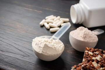 Two scoops of whey or soy protein powder, white capsules of amino acids, vitamins, creatine, protein chocolate bar, bodybuilding food supplements, sports nutrition on a dark wooden board