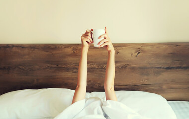 Cheerful lazy woman waking up after sleeping lying in soft comfortable bed showing empty cup coffee stretching her hands up from under the blanket in bedroom at home