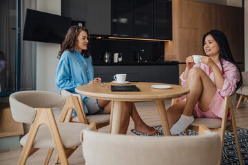 Fototapeta na wymiar Two beautiful women drinking coffee and talking while sitting in kitchen
