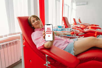 Young woman is holding a phone with the inscription Be a Hero Give Blood and making blood donation...