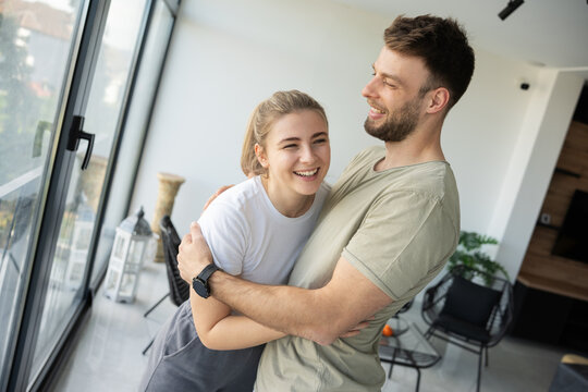 A married couple stands by the window at home, embracing each other in a tender moment of love and connection.