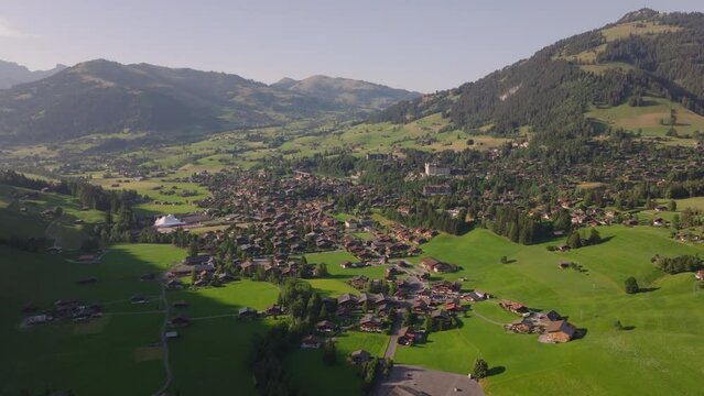 Fly above wide mountain valley. Typical houses and chalets in alpine town surrounded by green meadows and hills. Gstaad, Switzerland