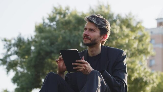 Young guy browsing tablet in garden. Thoughtful businessman decide to invest much money to crypto money. Man using mini computer researching news outdoor. Relaxing student reading ebook.